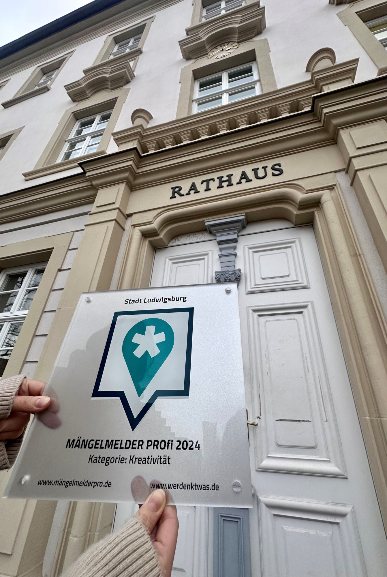 Die Award-Plakette zur Auszeichnung "Mängelmelder Profi 2024" wird vor der Rathaustür der Stadt Ludwigsburg hochgehalten.